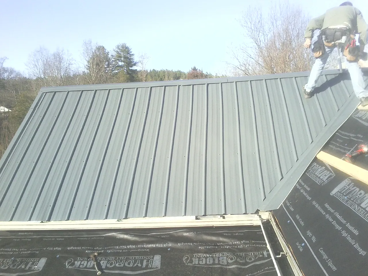 Crew installing dark green standing seam panels over synthetic underlayment in Coloma